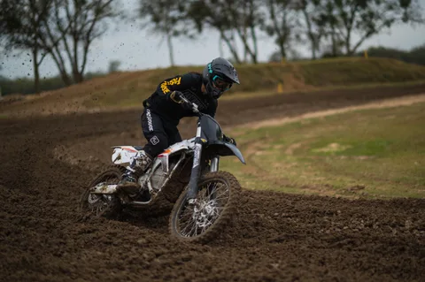 Rider in black gear leaning a white motocross bike into a rutted Creekside corner